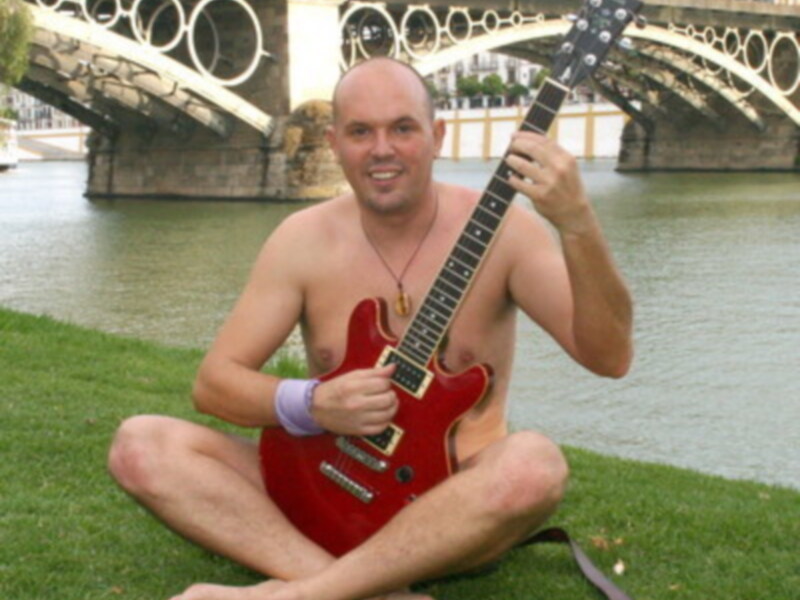 Un hombre sin camisa sostiene una guitarra eléctrica roja sentado en el césped junto a un río con un puente en el fondo.