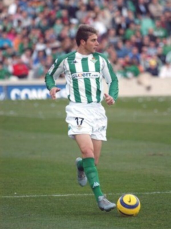 Un jugador de fútbol con una camiseta verde y blanca, marcada con el número 17, corre hacia un balón amarillo y azul en un campo de fútbol. La grada está llena de espectadores, y el césped es verde y bien mantenido.