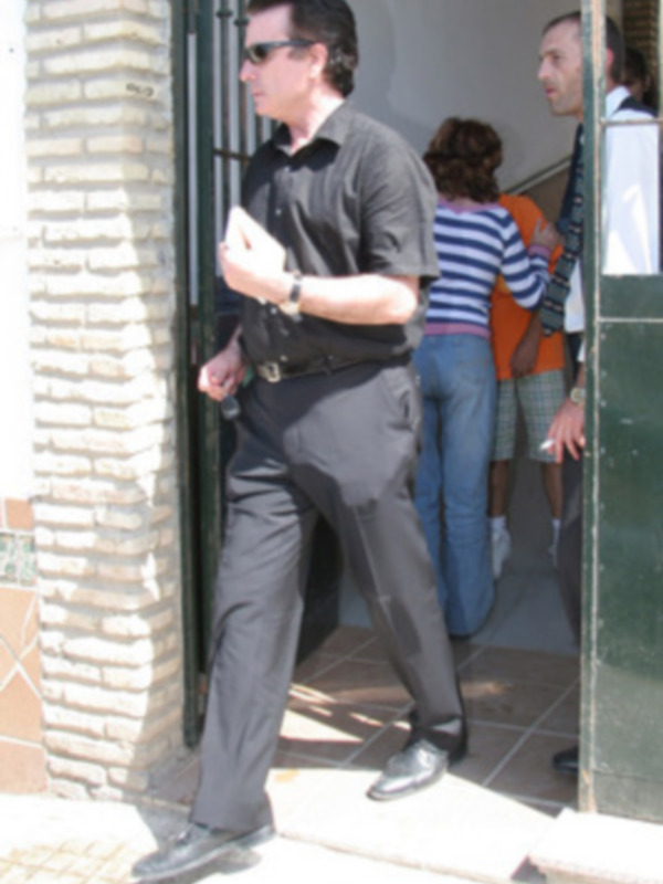 Un hombre con camisa negra y pantalones de vestir, caminando por una calle. Detrás, dos personas conversan en el fondo. La imagen muestra un ambiente urbano con una pared de ladrillos y una puerta metálica.