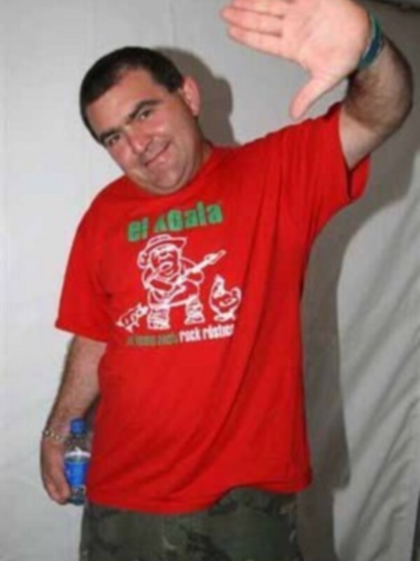 Un hombre con una camiseta roja que dice "El Gato" y pantalones de camuflaje, posando con una botella azul en la mano izquierda.