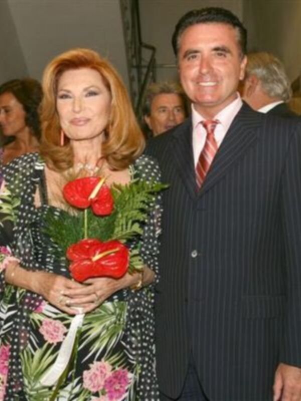 Una pareja posa juntos, ella sostiene una flor roja con hojas verdes y él lleva una camisa con corbata roja y una chaqueta de traje. Ambos parecen estar en un evento formal.