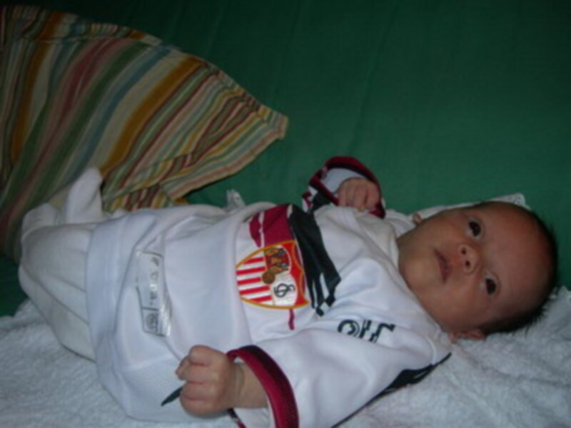Un bebé recién nacido está tumbado boca arriba, con una camiseta de fútbol del Sevilla FC sobre la ropa interior. El bebé tiene los ojos abiertos y parece estar dormido. Detrás de él, se pueden ver una almohada con rayas multicolores y un fondo verde.