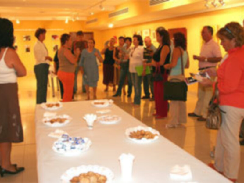 Una sala llena de personas observando una exposición. La mesa central tiene varios platos con diferentes objetos, posiblemente artefactos históricos. La gente está interesada y discutiendo entre sí.