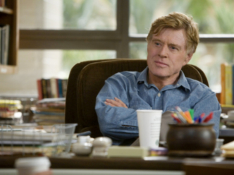 Un hombre sentado en una silla de oficina con las manos cruzadas, frente a una mesa con libros y un tazón de plumas.