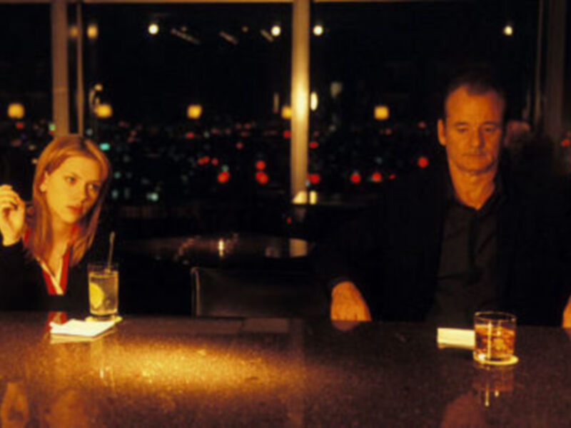 Una mujer y un hombre están sentados en una mesa de cristal, disfrutando de bebidas alcohólicas. La imagen está iluminada con luces brillantes en el fondo, creando un ambiente romántico.