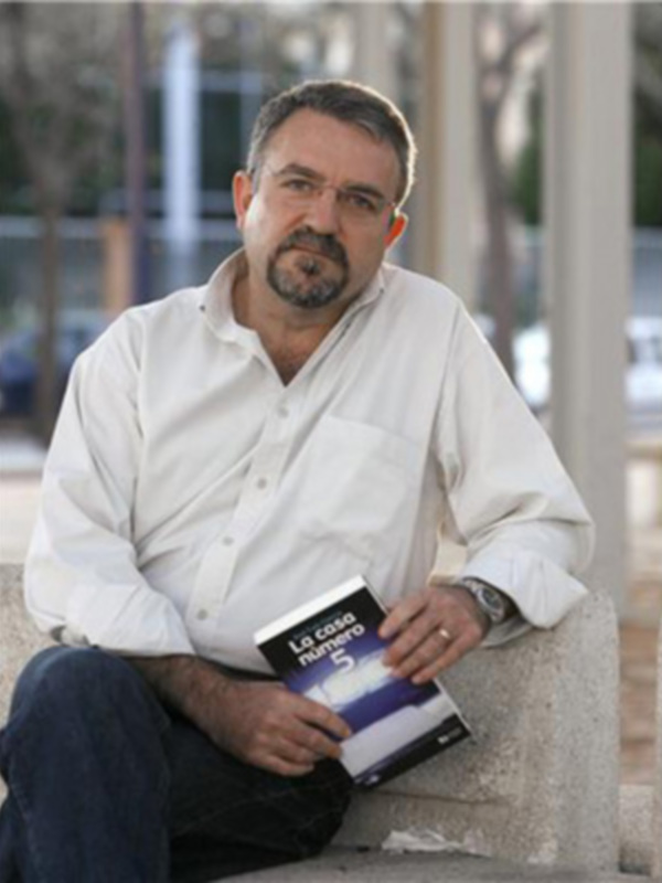 Un hombre con barba y cabello gris, vistiendo una camisa blanca, sostiene un libro titulado "La Casa Numerada 5". Se encuentra sentado en una banca de piedra, con un fondo urbano borroso.