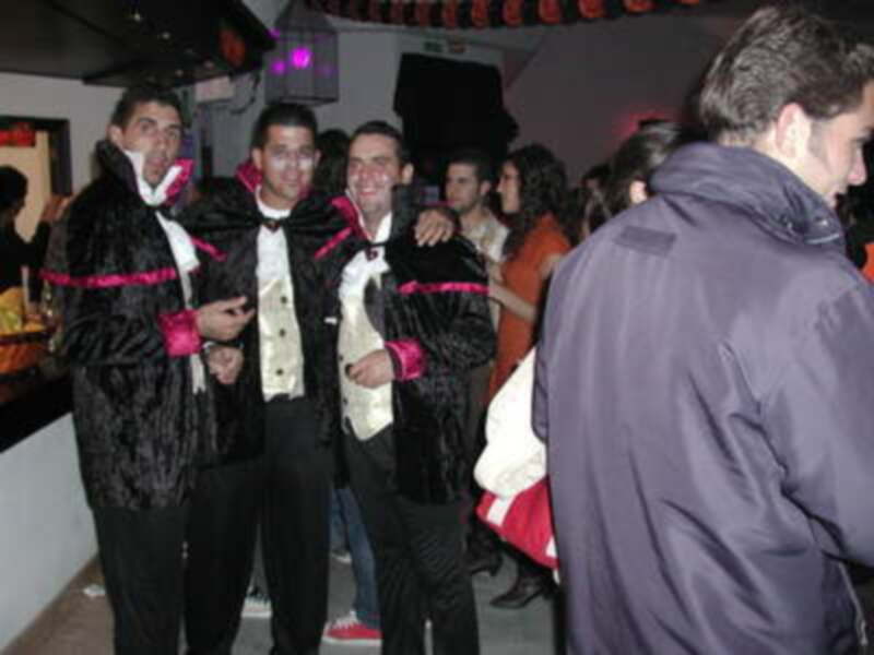 Dos jóvenes vestidos con trajes de gala se encuentran en una fiesta, rodeados por otros invitados. El ambiente es festivo y la iluminación es brillante, con luces rojas y blancas en el fondo.