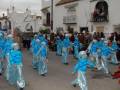 Desfile de carnaval en una calle con participantes disfrazados de personas con trajes azules. Edificio blanco en el fondo.