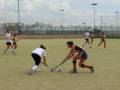 Jugadoras de hockey en acción sobre un campo de césped, con una torre de iluminación y estructuras metálicas al fondo.