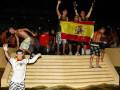 Un grupo de jóvenes celebrando en una playa nocturna, algunos saltan y otros levantan un bandera española.