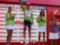 Un ciclista celebra en el podio con una bandera verde, rodeado por tres mujeres vestidas de verde y blanco. El fondo es rojo con anuncios de la Vuelta a España 2010.