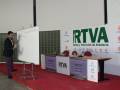 Un hombre en traje negro está presentando en un evento de RTVA, Radio y Televisión de Andalucía. La imagen muestra una mesa con un tablero blanco, un micrófono y varios logotipos de RTVA. El fondo es una pantalla con texto en verde y rojo, y hay una alfombra roja alrededor de la mesa.