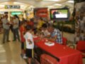 Un niño en una camiseta de fútbol roja y amarilla se prepara para firmar un autógrafo en una mesa roja con una bandera de fútbol detrás. En el fondo, se puede ver a otros niños y adultos en un espacio público con iluminación brillante.