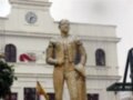 Estatua de un personaje histórico en una plaza con edificios y banderas al fondo.