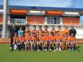 Equipo de hockey infantil posando en el estadio, con uniformes naranjas y azules, frente a una estructura deportiva con marcos de color naranja y azul.