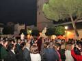 Una procesión nocturna con una imagen de la Virgen en el centro, rodeada por una multitud de personas. La escena se desarrolla frente a un edificio con una torre, y hay luces que iluminan la noche. El ambiente es festivo y religioso.