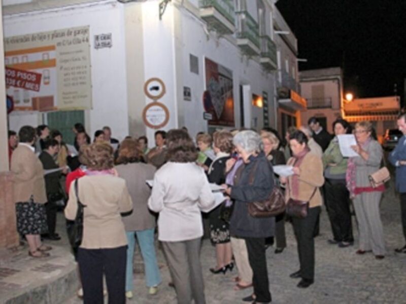 Una multitud de personas se reúne en una calle nocturna, frente a un edificio con carteles informativos. Algunos llevan bolsas y documentos, mientras otros observan el evento con interés. La escena transmite una sensación de expectativa y curiosidad entre los asistentes.