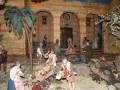 Un bello escenario navideño con figuras de personas y animales en una representación de la natividad. La escena muestra a personas con trajes antiguos, una palmera y un edificio de fondo.