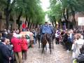 Personas observando a dos policías montados en caballos en una calle adoquinada, con árboles verdes y edificios al fondo.