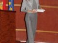 La Reina Letizia de España, con un elegante traje gris y una chaqueta con detalles en la parte superior, se encuentra frente a un escenario con banderas española y europea.