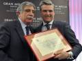 Dos hombres en trajes sostienen un diploma frente a una pared con el logo del Gran Meliá Hotel.