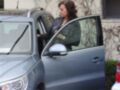 Una mujer está bajando del asiento trasero de un coche azul, con una chaqueta verde y zapatos negros. La imagen muestra un entorno urbano con árboles sin hojas y una pared de ladrillo.