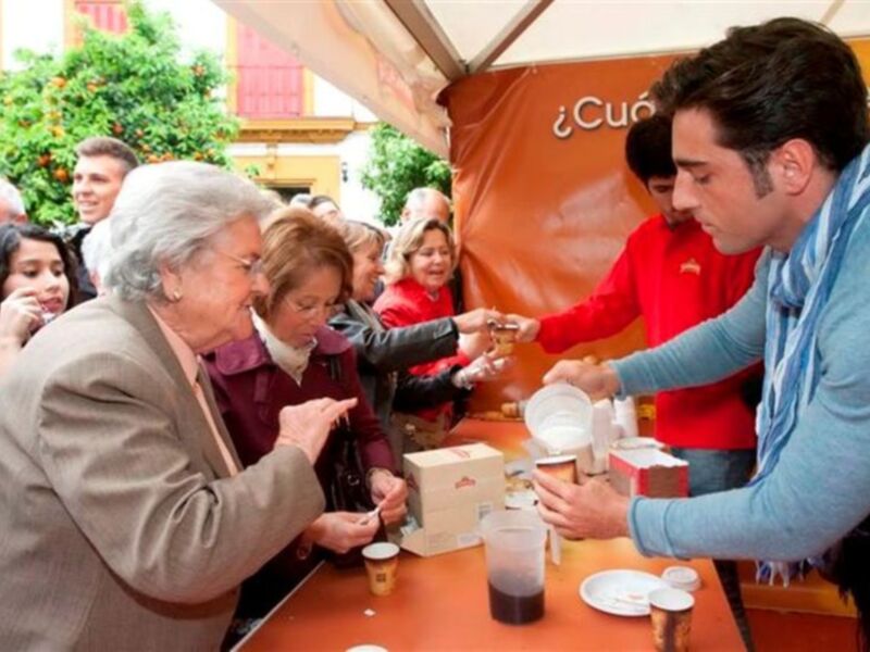 Un hombre en un chaqueta azul está sirviendo café a una mujer mayor. La imagen muestra una escena de servicio en un evento público, con varios asistentes y clientes. La gente está reunida alrededor de una mesa donde se sirven bebidas calientes, y el ambiente parece ser agradable.