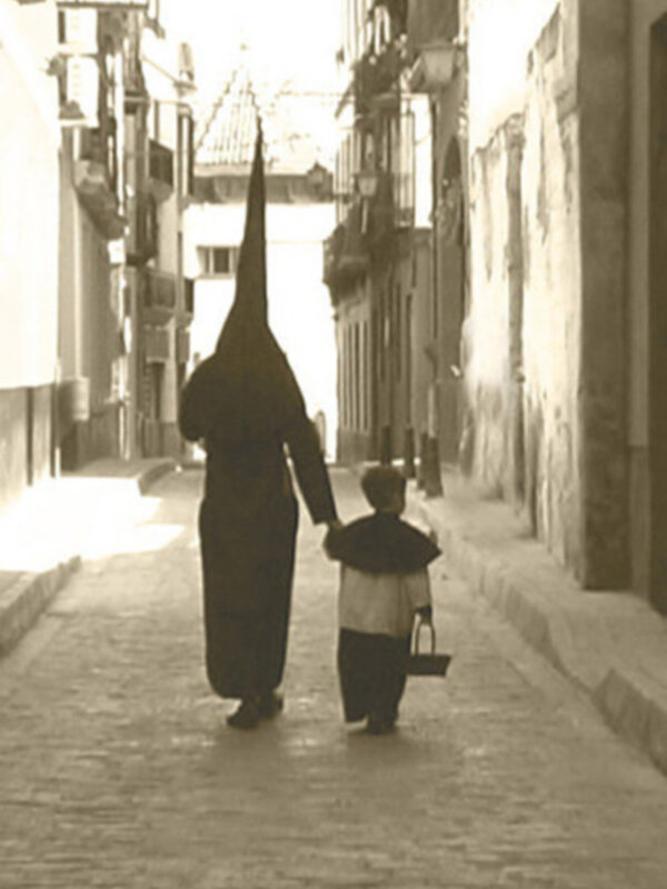 Una persona vestida con un traje tradicional de Semana Santa camina por una calle estrecha, seguida por un niño que lleva una cesta. La imagen es en blanco y negro, capturando el ambiente de la celebración religiosa.