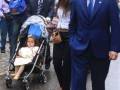 Una familia camina por la calle, con una niña en un cochecito y dos adultos. La imagen muestra a una mujer con una chaqueta blanca, un hombre en traje azul y una niña con zapatos de tacón. La escena parece ser en un día lluvioso, dado el aspecto mojado de la calle.