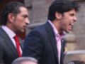 Dos hombres en trajes de negocios, uno con corbata roja y el otro con corbata rosa, posan juntos frente a un edificio con una chimenea en el fondo.