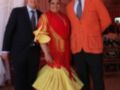Tres personas posan juntas, dos hombres en trajes y una mujer en un vestido flamenco. La imagen parece ser de una fiesta o evento formal, con cortinas y luces en el fondo.