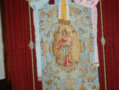 Un hermoso escudo religioso colgado en un fondo rojo, con una imagen de la Virgen María en el centro. La bandera está bordada y tiene detalles dorados, con un arco de oro en la parte superior.