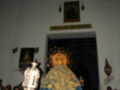 Una procesión religiosa en un templo. En el centro, se ve una imagen de la Virgen María con un manto azul y dorado. La capilla está decorada con una estatua de la Virgen en el altar y un cartel que dice "Capilla de San Cristóbal". La multitud está reunida para observar la procesión.