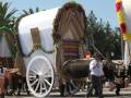 Imagen de un carro blanco adornado con flores y guirnaldas, tirado por dos bueyes. Un hombre camina al frente del carro, y una mujer con falda flamenca lo sigue. El fondo muestra un ambiente festivo con palmeras y otros ornamentos decorativos.