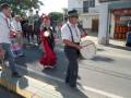 Person playing a drum in a street parade, wearing traditional attire and surrounded by other participants.