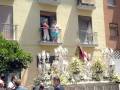 Una procesión religiosa con una imagen de Cristo y flores blancas, mientras personas observan desde balcones.