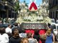Un grupo de personas camina por una calle con un carro cargado de flores blancas y una figura envuelta en velo rojo. El cielo es claro y el ambiente parece ser una procesión religiosa o festiva.