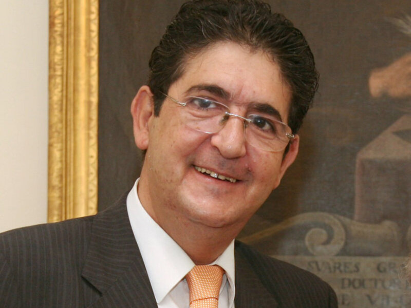 Un hombre con cabello oscuro y barba, lleva un traje negro con una camisa blanca y corbata naranja. Está sonriendo y parece estar en una situación formal, posiblemente un evento o reunión.