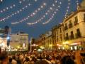 Festival nocturno en una plaza con farolillos colgados del cielo y multitudes animadas.