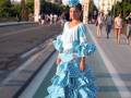 Una mujer vestida con un traje de flamenca azul con lunares blancos y un tocado similar, camina por una calle con una arquería de entrada a un parque en el fondo.