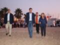 Un grupo de personas caminando en la playa, con palmeras y una estructura de luces al fondo.