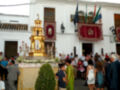 Procesión religiosa con estandartes y altares portátiles en una plaza de pueblo andaluz.