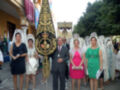 Una procesión religiosa con una imagen central de un trono dorado, acompañada por mujeres vestidas en trajes tradicionales y hombres con corbatas. La escena está ambientada en una calle con edificios de estilo mediterráneo y banderas ondeando.