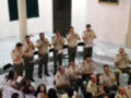 Un grupo de músicos militares toca instrumentos de viento en una sala con paredes blancas y ventanas. Los músicos están vestidos con uniformes militares, y la sala parece ser parte de un edificio histórico o institucional.