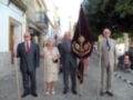 Una imagen de una procesión religiosa con cuatro personas vestidas formalmente, dos hombres y una mujer, sosteniendo un bastón y una bandera con emblemas. La escena está rodeada de edificios blancos y una iglesia en el fondo, con un parque o jardín al lado.