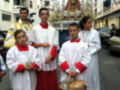 Una procesión religiosa en una calle con personas vestidas de blanco y rojo, sosteniendo un cesto. En el fondo, una imagen religiosa en una carroza.