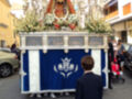 Una procesión religiosa con una imagen de la Virgen en un carro, rodeada por flores y decoraciones. La imagen se realiza frente a edificios urbanos, con personas observando la procesión.