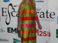 Una mujer con un vestido largo de colores brillantes, posa frente a una pared con logos de empresas.