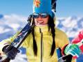 Excited skier in vibrant yellow suit and colorful beanie, holding skis and poles against a snowy mountain backdrop.