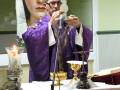 En un altar, un sacerdote vestido de morado sostiene una imagen religiosa mientras realiza una ceremonia. Una vela encendida y un cáliz dorado se encuentran en el centro, rodeados por elementos de decoración tradicional.